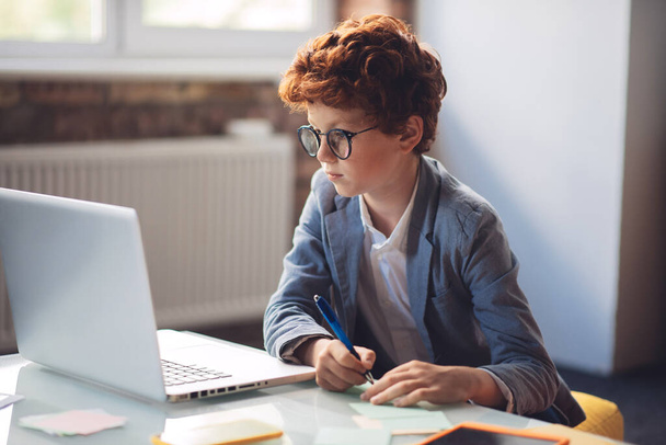 Red-haired boy in sitting at the laptop and looking serious - Photo, image