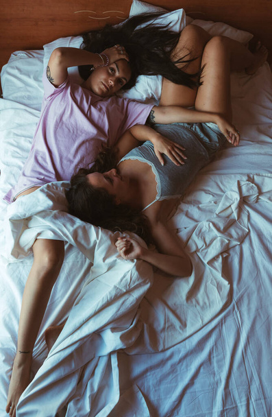 Cena suave de duas jovens mulheres deitadas em uma cama com lençóis brancos abraçando com uma expressão pensativa. - Foto, Imagem