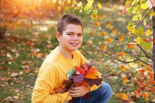 Školák v parku zblízka. Usmívající se pohledný kavkazský teenager ve žluté košili s dlouhým rukávem a modrých džínách, sedící v parku s kyticí podzimních listů. Podzimní koncept. Kopírovat prostor  - Fotografie, Obrázek