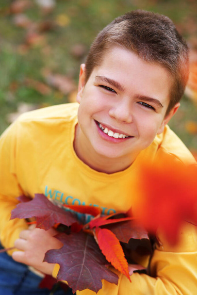 Smiling handsome caucasian teenager boy close-up in a yellow long sleeve shirt and blue jeans sitting in a park with a bouquet of autumn leaves. - Photo, Image