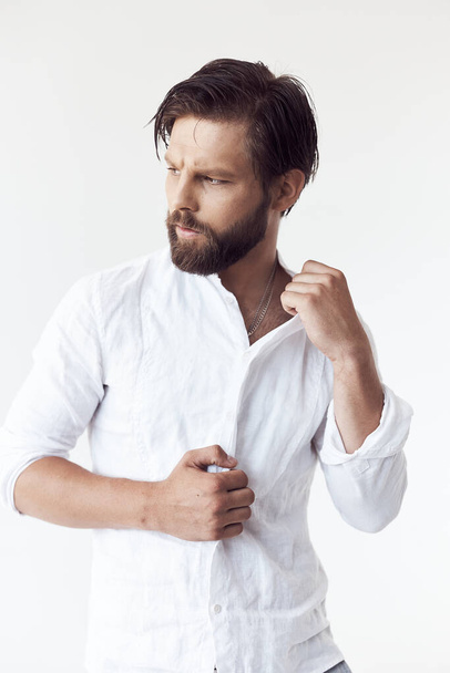 portrait photo of a handsome bearded man with brown hair on white background, he is wearing a white linen shirt, looks away and touches his shirt - 写真・画像