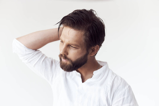 high key portrait photo on white cyclorama of a handsome bearded man with brown hair and eyes, he is wearing a white linen shirt, he holds his hand behind his head and looks away - Фото, зображення