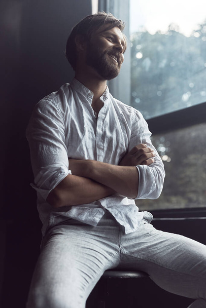 photo of a handsome bearded man with brown hair who sits on a chair in the studio, he is wearing a white linen shirt and pants, smiles and looks away - Zdjęcie, obraz