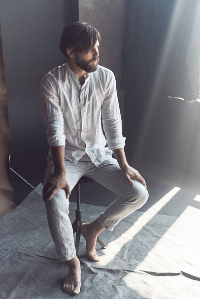 full-length photo of a handsome bearded man with brown hair who sits on a chair in the studio with a ray of sunshine, he is wearing a white linen shirt and pants and looks away - Zdjęcie, obraz