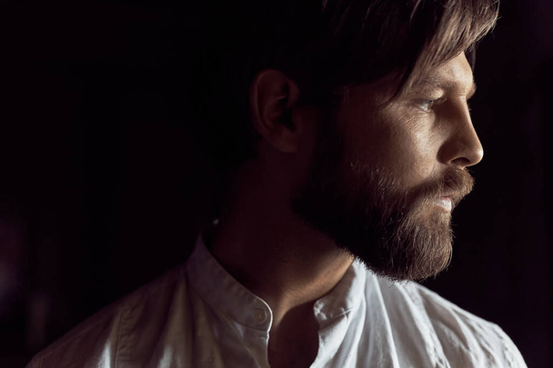 low key portrait photo of a handsome bearded man with brown hair wearing a white linen shirt, he looks in right direction - Fotoğraf, Görsel