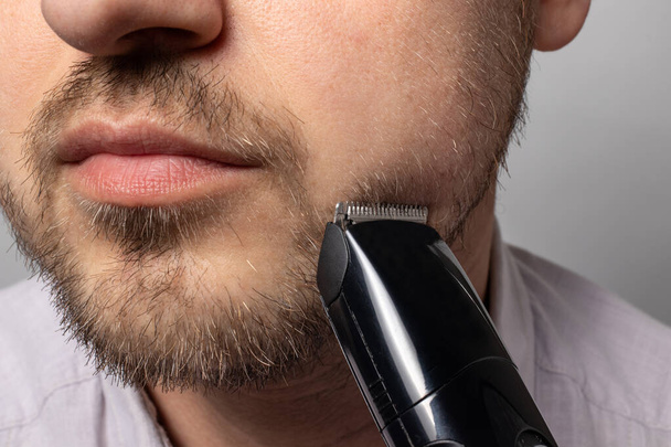 A man shaves his beard with a trimmer razor. Modeling beard, masculine style, facial hair care, morning routines in the bathroom. - 写真・画像