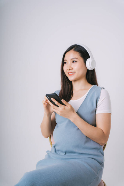 Krásná těhotná asijské žena poslouchat hudbu s sluchátkem a pomocí smartphone izolovat přes bílé pozadí. - Fotografie, Obrázek