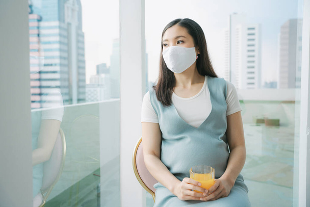 Pamuk maskeli güzel hamile bir Asyalı kadın elinde bir bardak portakal suyuyla sandalyede oturuyor. Evde karantina. - Fotoğraf, Görsel