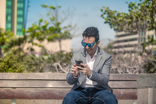 Позитивный взрослый мужчина-предприниматель в шикарном наряде и солнечных очках сидит на скамейке на городской улице и счастливо улыбается, получая отличные новости на смартфоне - Фото, изображение
