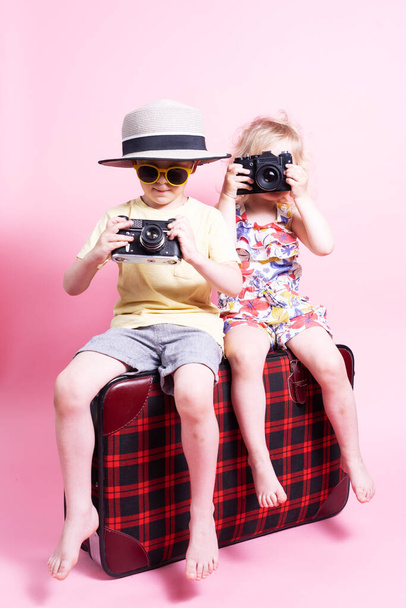 Cestování: Děti malý chlapec a dívka hrát v turisty, sedět na velký červený kufr a fotit. Studio portrét na růžovém pozadí. - Fotografie, Obrázek