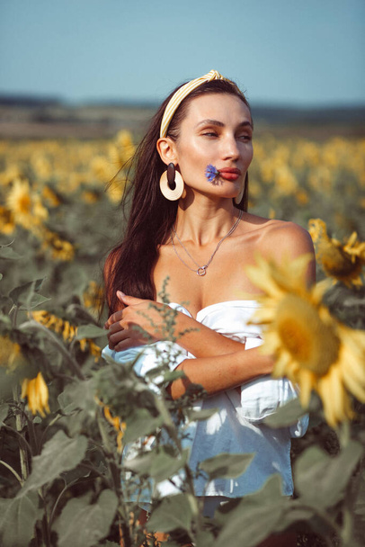 a young beautiful girl, with an expressive face and beautiful lips, she stands among a bright yellow field of sunflowers, she has a beautiful hat and a white shirt on her naked body - Foto, Bild