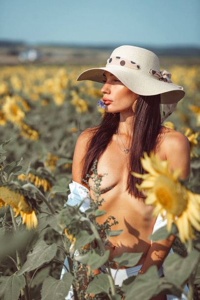 a young beautiful girl, with an expressive face and beautiful lips, she stands among a bright yellow field of sunflowers, she has a beautiful hat and a white shirt on her naked body - Foto, Imagen