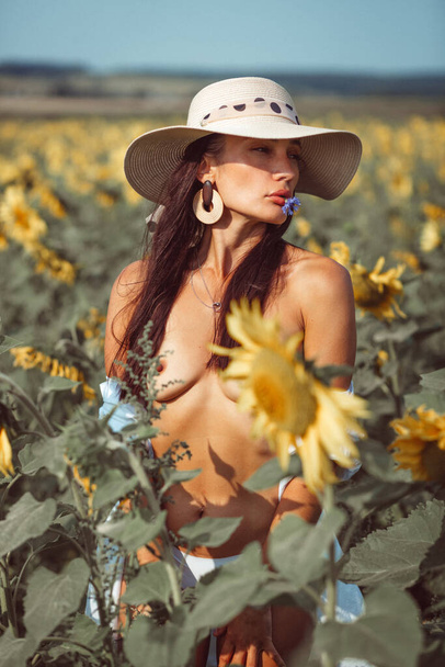 a young beautiful girl, with an expressive face and beautiful lips, she stands among a bright yellow field of sunflowers, she has a beautiful hat and a white shirt on her naked body - Valokuva, kuva