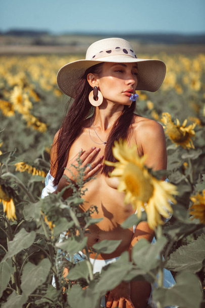 a young beautiful girl, with an expressive face and beautiful lips, she stands among a bright yellow field of sunflowers, she has a beautiful hat and a white shirt on her naked body - Photo, Image