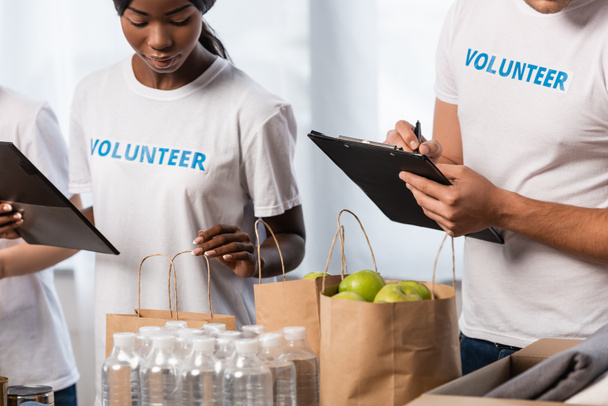 Selective focus of multiethnic volunteers with clipboards standing near packages with food and bottles of water  - Photo, Image