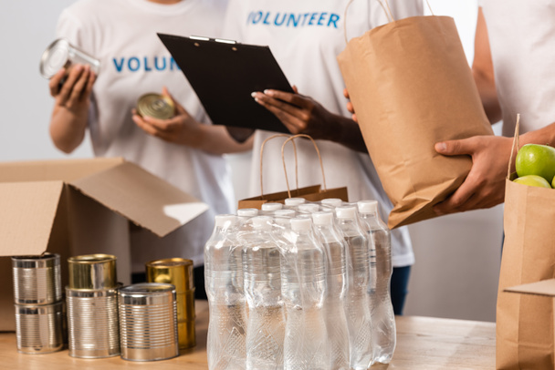 Vue recadrée de bénévoles multiethniques avec presse-papiers debout près des bouteilles d'eau et des emballages avec de la nourriture dans un centre caritatif   - Photo, image