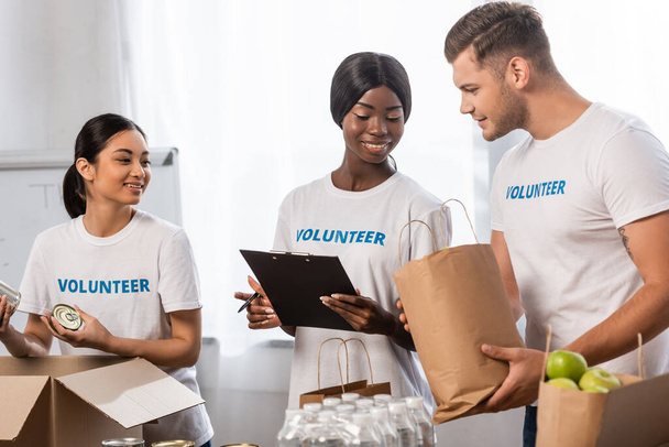 Multiculturele vrijwilligers houden pakketten met voedsel in de buurt van Afrikaanse Amerikaanse vrouw met klembord in een liefdadigheidscentrum   - Foto, afbeelding