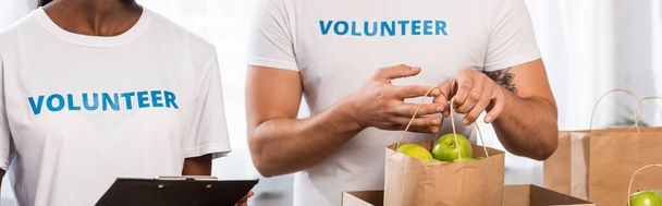 Panoramic crop of multiethnic volunteers holding clipboard and paper bag with apples in charity center  - 写真・画像