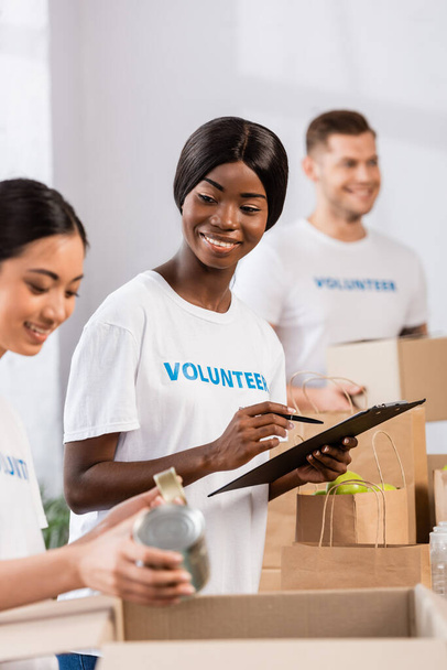 Selectieve focus van Afrikaanse Amerikaanse vrijwilliger met klembord in de buurt van aziatische vrouw met blikken in liefdadigheidscentrum  - Foto, afbeelding