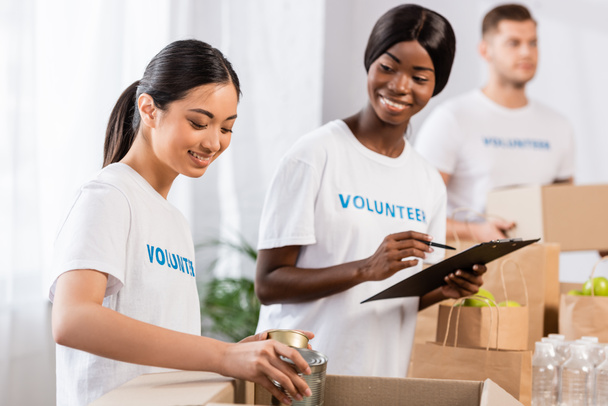 Selectieve focus van aziatische vrijwilliger zetten blikken in doos in de buurt van Afrikaanse Amerikaanse vrouw met klembord in liefdadigheidscentrum  - Foto, afbeelding