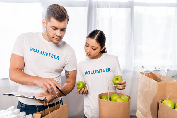 Enfoque selectivo de voluntarios multiculturales con portapapeles y manzanas de pie cerca de paquetes en el centro de caridad  - Foto, Imagen