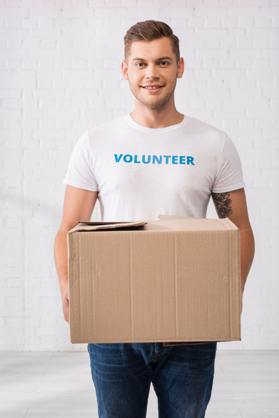 Volunteer in white t-shirt holding cardboard package in charity center  - Foto, afbeelding