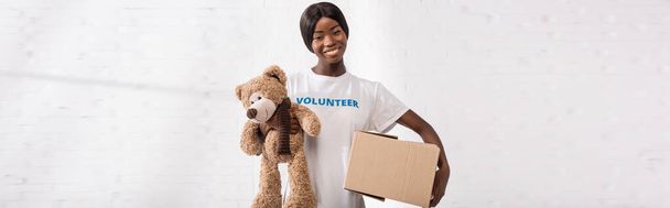 Pozioma uprawa afrykańskiego wolontariusza z miękką zabawką i kartonowym opakowaniem patrzącym w kamerę  - Zdjęcie, obraz
