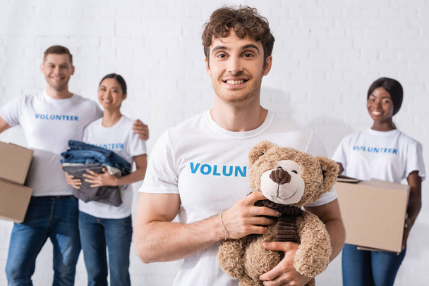 Selective focus of man holding soft toy near multicultural volunteers with packages in charity center  - Photo, Image