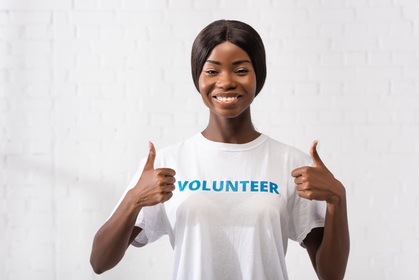 Donna afroamericana in t-shirt con lettering volontario che mostra i pollici in alto  - Foto, immagini