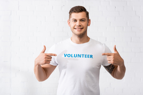 Young volunteer pointing at lettering on white t-shirt and looking at camera  - Фото, зображення