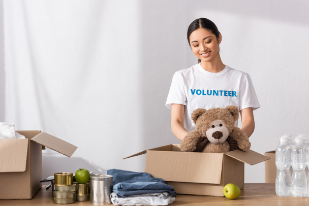 Asian volunteer putting soft toy in carton package near clothes and tin cans in charity center  - 写真・画像