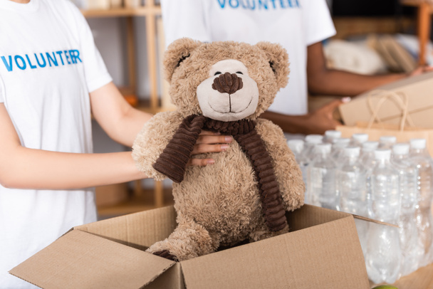 Vista cortada de voluntário colocando brinquedo macio em caixa de papelão perto de pacotes com garrafas de água no centro de caridade  - Foto, Imagem