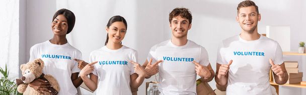 Горизонтальный урожай молодых мультиэтнических волонтеров, указывающих пальцами на надписи на футболках и показывающих большие пальцы в благотворительном центре  - Фото, изображение