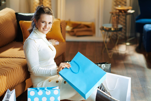 happy stylish woman in white sweater and skirt sitting near sofa with shopping bags at modern home in sunny winter day. - Foto, immagini