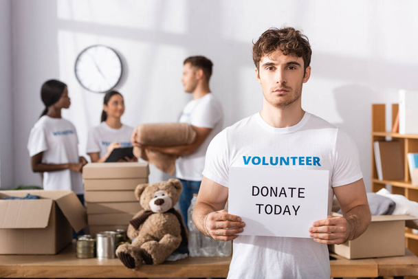 Selectieve focus van vrijwilliger holding card met doneren vandaag belettering met multi-etnische mensen op de achtergrond in het liefdadigheidscentrum  - Foto, afbeelding
