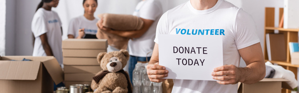Horizontal image of volunteer holding card with donate today lettering near multiethnic people in charity center at background  - Photo, Image