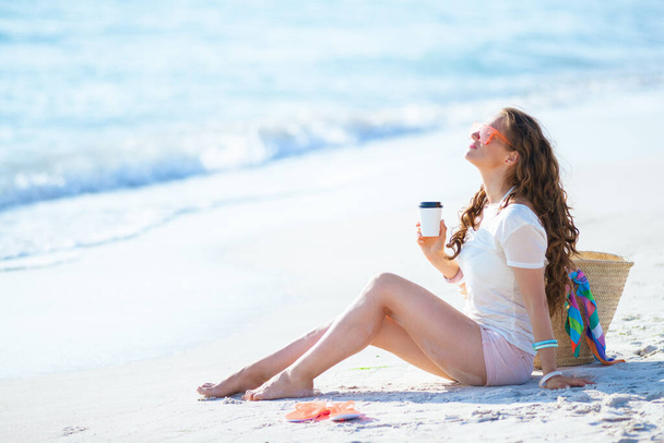 relaxed young woman in white t-shirt and pink shorts on the ocean coast drinking coffee while sitting. - Фото, изображение