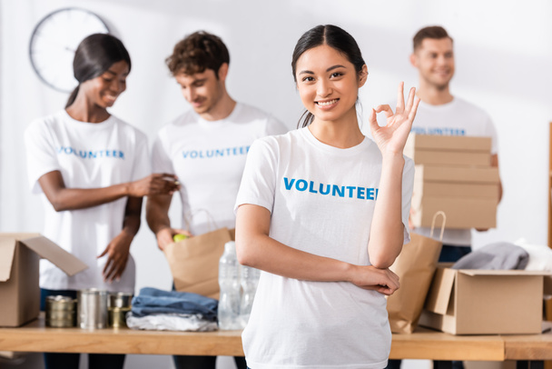 Foco seletivo de voluntário asiático mostrando gesto ok perto de doações e pessoas multiétnicas no centro de caridade em segundo plano  - Foto, Imagem