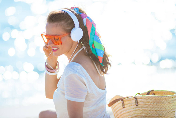 smiling trendy woman in white t-shirt and pink shorts listening to the music with headphones on the ocean coast. - Foto, imagen