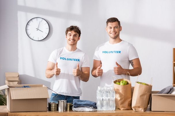Voluntarios mostrando pulgares hacia arriba cerca de paquetes y ropa en la mesa en el centro de caridad  - Foto, imagen