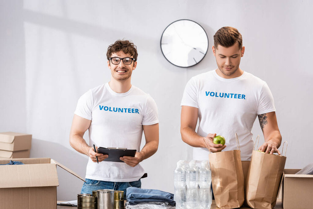 Selective focus of volunteers with clipboard and apple standing near donations on table  - Foto, immagini
