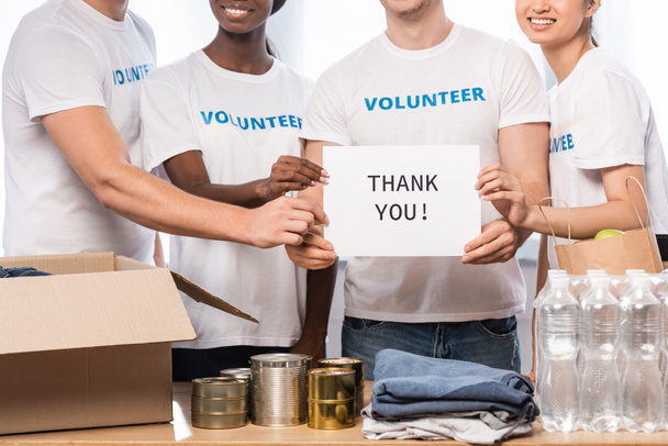 Cropped view of multiethnic volunteers holding card with thank you lettering and donations in charity center  - Photo, Image