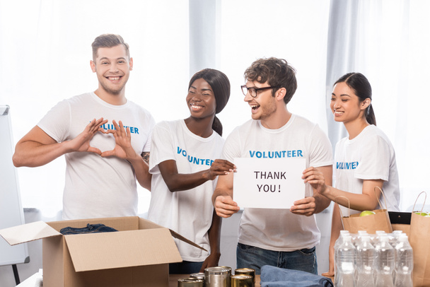 Selektiver Fokus der Freiwilligen zeigt Herzzeichen in der Nähe von lachenden multiethnischen Menschen mit Schriftzug Danke auf Karte und Spenden  - Foto, Bild