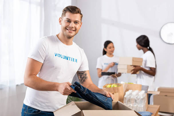 Selectieve focus van vrijwilliger die naar de camera kijkt terwijl hij kleding in een kartonnen doos in een liefdadigheidscentrum stopt  - Foto, afbeelding