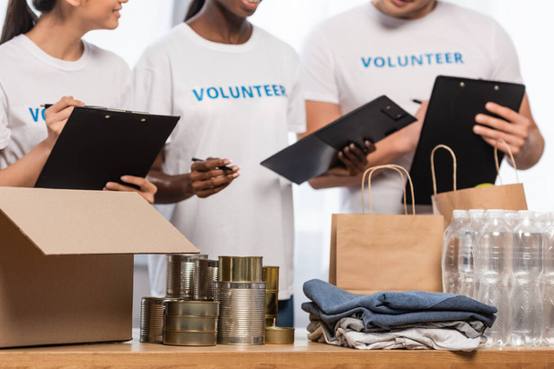 Vágott kilátás többnemzetiségű önkéntesek vágólapokkal közel ón dobozok, ruhák és csomagok jótékonysági központ  - Fotó, kép
