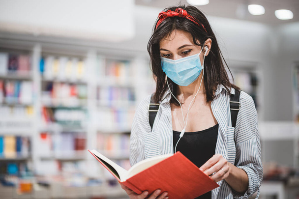 Tıp maskesi takan ve Coronavirs salgını sırasında kütüphanede kitap okuyan çekici bir bayan öğrenci. Not defterinden okuyan tatlı bir kız. Eğitim, lise, üniversite, öğrenim ve insanlar kavramı - Fotoğraf, Görsel