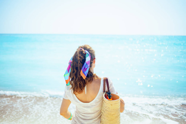 Seen from behind trendy woman in white t-shirt with beach straw bag on the ocean coast. - Photo, Image
