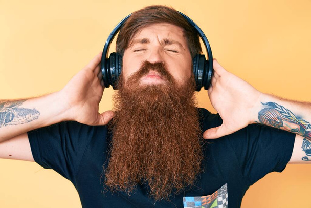 Handsome young red head man with long beard listening to music using headphones looking at the camera blowing a kiss being lovely and sexy. love expression.  - Fotó, kép