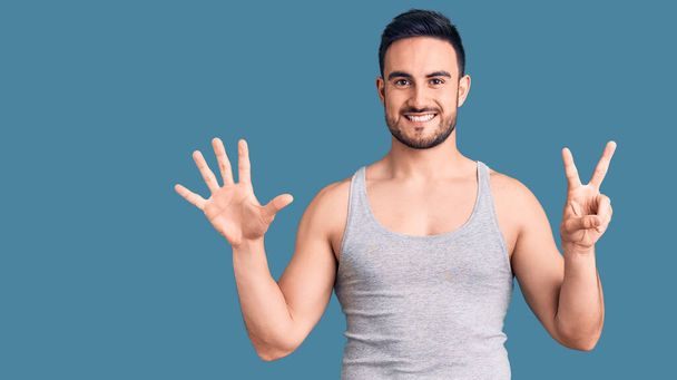 Junger gutaussehender Mann in Badebekleidung und ärmellosem T-Shirt zeigt mit Finger Nummer sieben nach oben und lächelt dabei selbstbewusst und glücklich.  - Foto, Bild