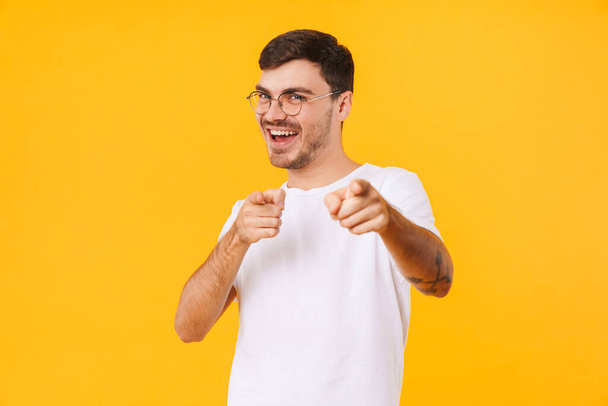 Photo of joyful young man in eyeglasses pointing fingers at camera and smiling isolated over yellow background - Zdjęcie, obraz